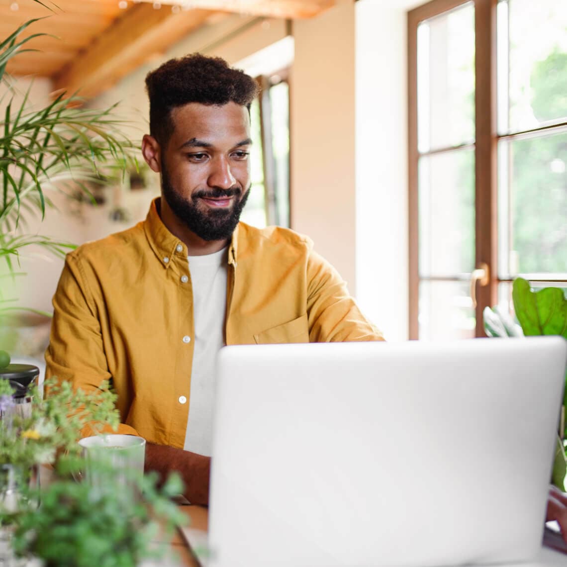 man in yellow button up shirt sitting at a laptop by a window